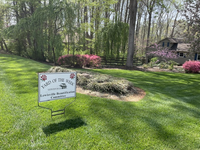 The "Yard of the Week" lawn sign stands in a healthy green lawn in front of a wooded area. A tan home sits between the two, by pink and purple flowering bushes.
