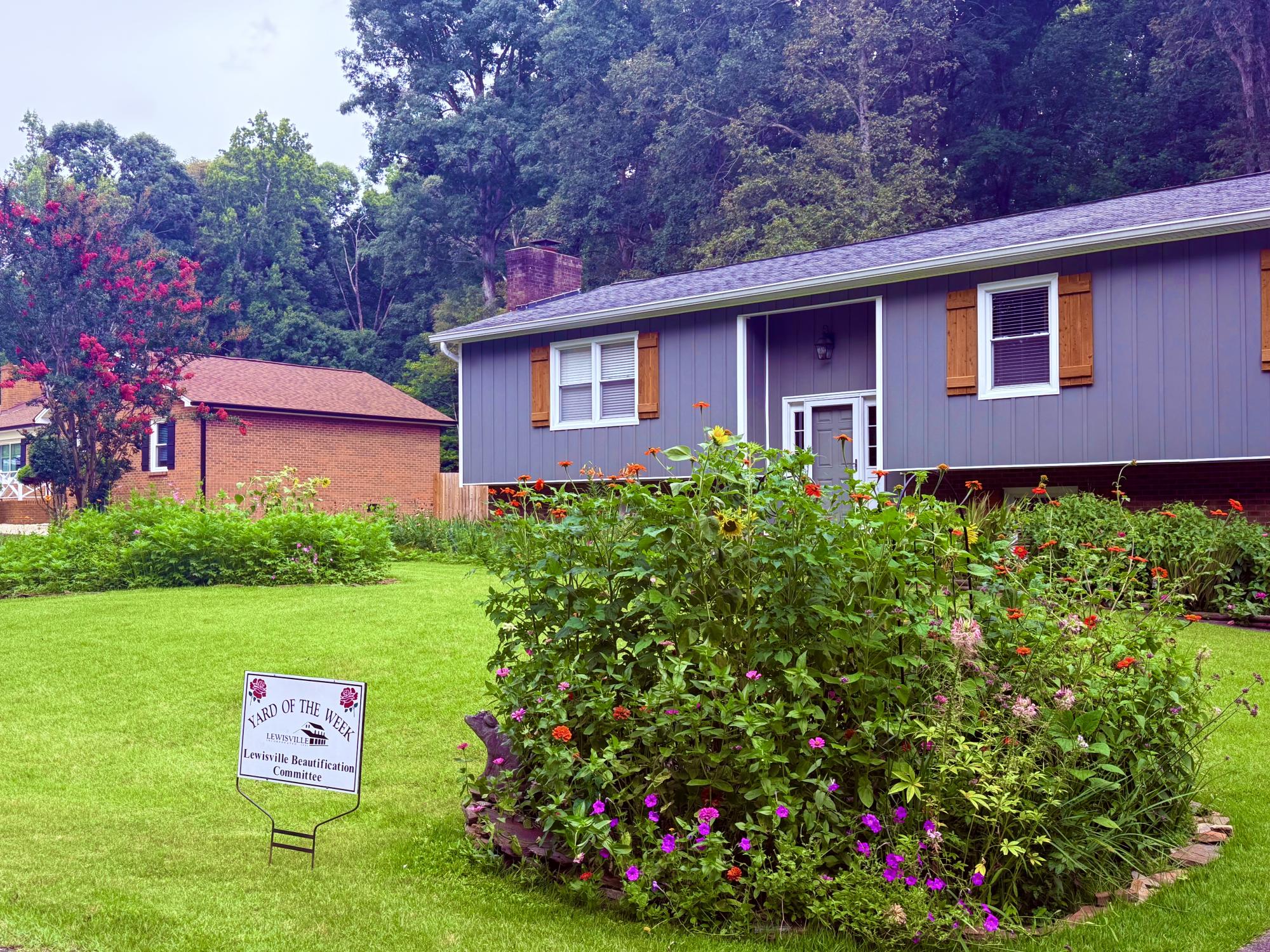 A medium gray house with wooden shutters sits surrounded by a manicured lawn with beautiful bushes.
