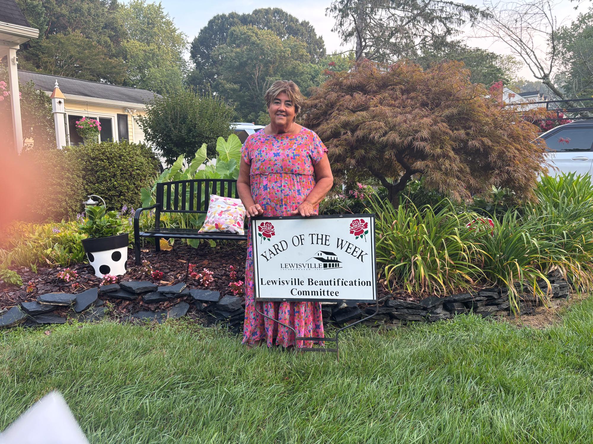 A woman in a pink dress stands by the Yard of the Week sign in front of her beautiful landscaping.