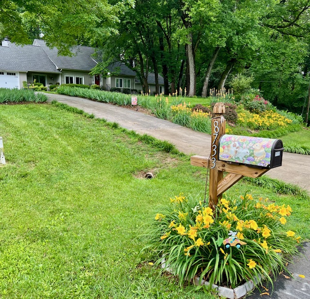 A colorful mailbox stands in a flower garden at the end of a manicured lawn with islands of flowers