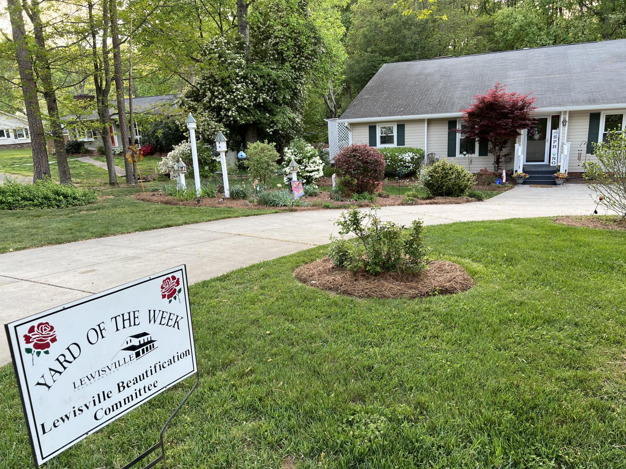 The Yard of the Week lawn sign is in front of a quaint white home with a a beautiful lawn, full of birdhouses and bushes sits tucked in a lush wooded area.
