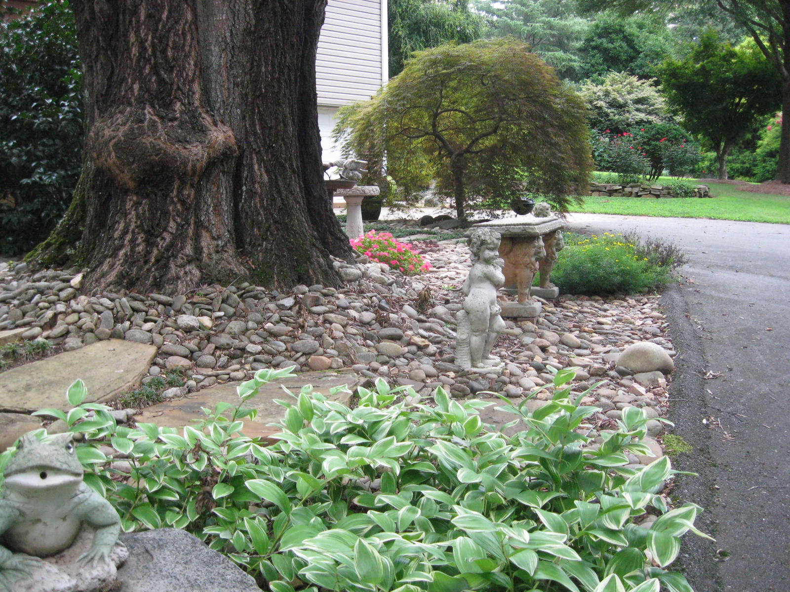 A stone garden has colorful flowers and plants around a large tree with two mini statues and a bench