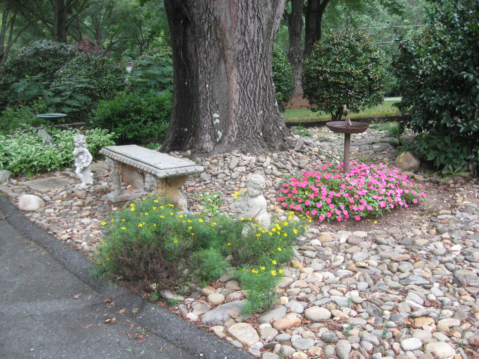 A stone garden has colorful flowers and plants around a large tree with two mini statues and a bench