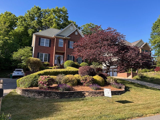 A multi-story brick house sits on a hill surrounded by beautiful landscaping and a colorful tree.